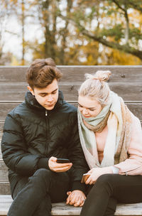 Young couple using smart phone while sitting on bench at park during autumn