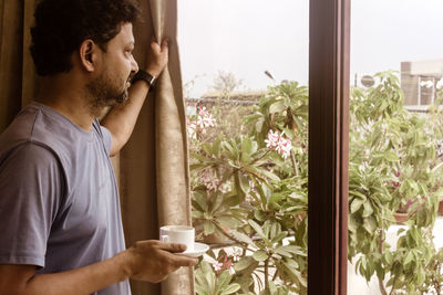 Man holding coffee cup while looking through window at home