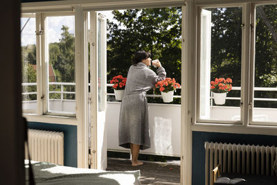 Rear view of woman enjoying tea while leaning on railing in balcony seen through doorway