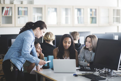 Mature teacher assisting female students using laptop in computer lab at high school