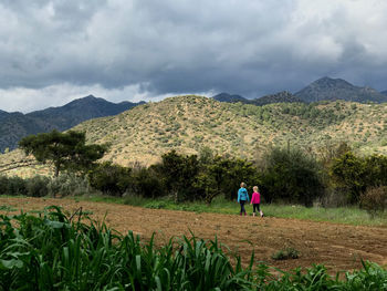 Rear view of sibling walking on field against mountains and sky