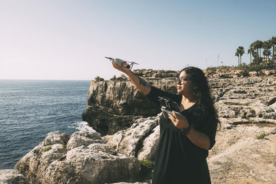 Happy woman with drone looking to the side black dress  by the sea sunny day. technological concept