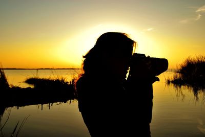 Silhouette woman photographing from dslr camera by lake against sky during sunset