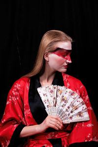 Young female model holding hand fan while sitting against black curtain