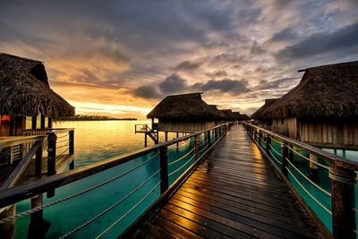 Pier and stilt houses over sea against cloudy sky during sunset