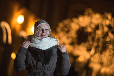 Cheerful young woman standing against illuminated christmas lights at night