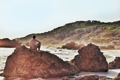 Rear view of man sitting on rock by sea against clear sky