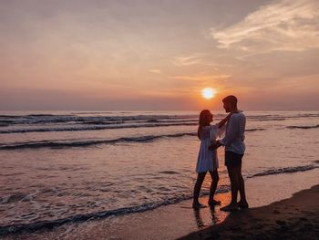 Rear view of friends standing at beach against sky during sunset