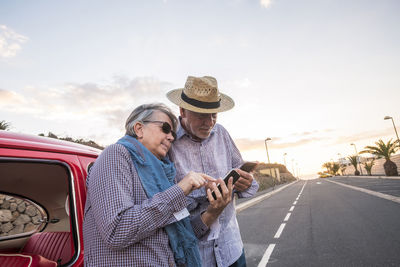 Senior couple using mobile phone while standing by car against sky
