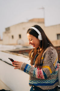 Smiling woman wearing headphones and using phone on building terrace