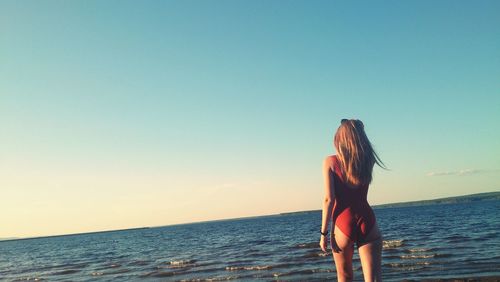 Rear view of sensuous woman in swimsuit walking on shore against blue sky