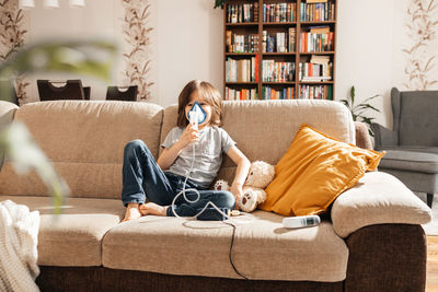 A little boy makes an inhalation at home sitting on the couch using a nebulizer and a mask. 