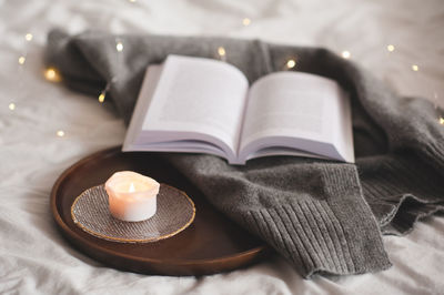 Scented burning candle on plate with knitted textile and open paper book on wooden tray in bed home