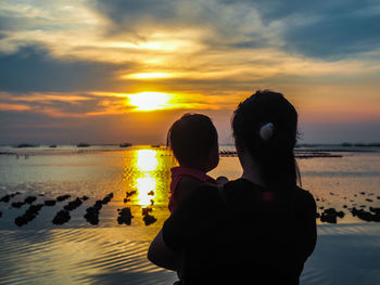 Rear view of woman with daughter at beach against sky during sunset