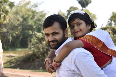 Portrait of smiling man piggybacking daughter against trees