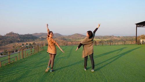 Young female friends with arms outstretched standing on grassy field against sky