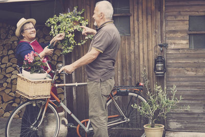 Senior couple standing with potted plant in bicycle basket against house