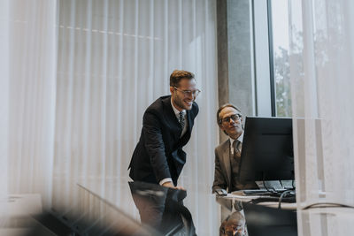 Smiling male business colleagues discussing over computer at desk in office