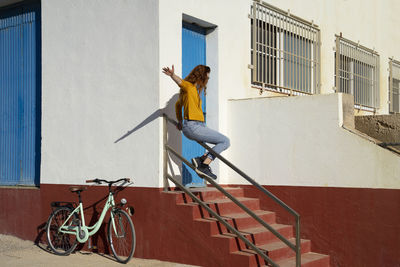Young woman sliding on railing by white built structure during sunny day