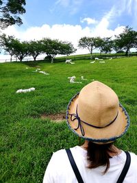 Rear view of mid adult woman wearing hat looking at goats on grassy field against cloudy sky