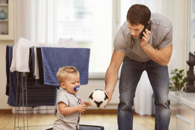 Father using mobile phone while giving ball to baby boy at home