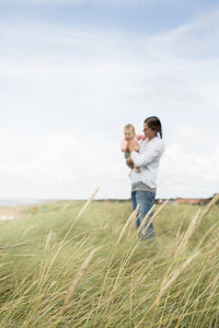 Woman carrying baby while standing on grassy field against sky