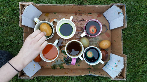 Cropped hand of woman over cardboard box with dye and easter eggs in cups