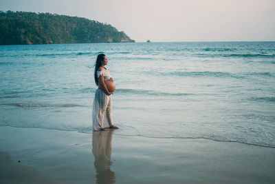 Side view of pregnant woman standing on shore at beach against sky