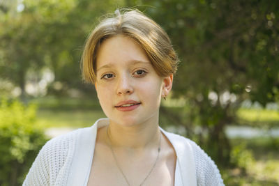 Close-up portrait of a red-haired teenage girl in the park against the backdrop of foliage.