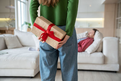 Gift box with ribbon for holiday. woman holding present behind back going to man who lying on couch