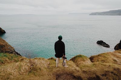 Rear view of man with camera standing at shore against cloudy sky