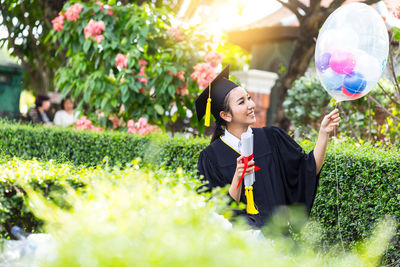 Smiling young woman in graduation gown holding balloon while standing by plants