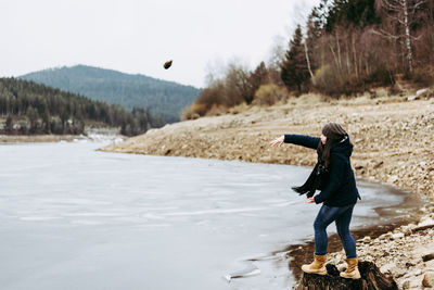 Side view full length of woman throwing stone while standing at riverbank during winter