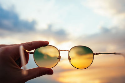 Hand holds metal round sunglasses through which you can see sea sunset in blue and orange colors