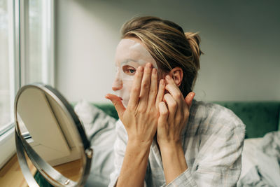 A woman applies a moisturizing face mask while sitting in front of a mirror in the bedroom.