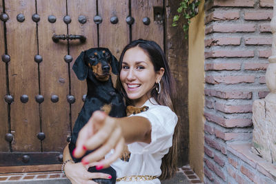 Portrait of happy woman gesturing while holding dog at house doorway