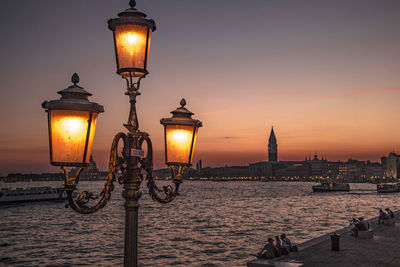 Illuminated street light by sea against sky at sunset