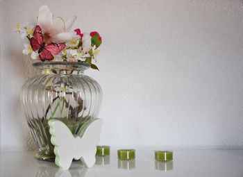Close-up of flowers in vase with artificial butterflies by tea lights on table against white wall