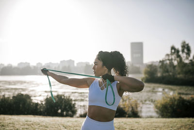 Female athlete stretching resistance band while exercising in public park against clear sky