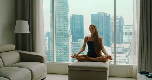 Woman meditating by the window with view on modern buildings skyscrapers of a big city. malaysia.