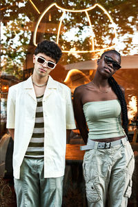 Young diverse man and woman in trendy clothes and sunglasses standing against reflective glass wall in daytime on city street