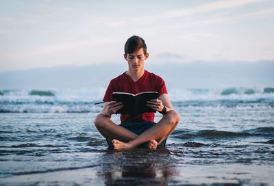 Full length of young man reading book at beach against sky