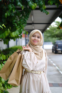 Young woman standing against shopping mall after shopping while holding paper bag 