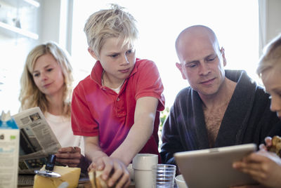 Man and sons using digital tablet while mother reading newspaper at dining table
