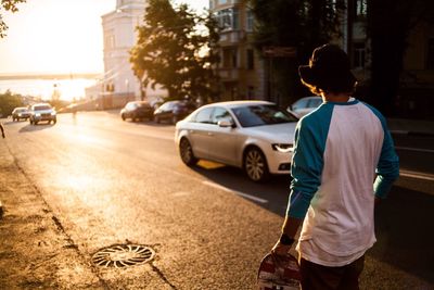 Rear view of man carrying skateboard while walking on road during sunset