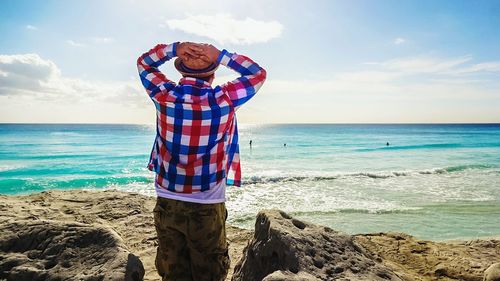 Rear view of man with arms raised standing at beach against sky on sunny day