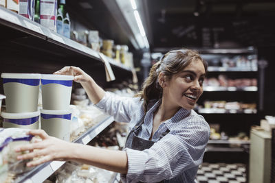 Young female owner looking over shoulder while arranging product on rack at delicatessen shop