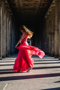 Full length side view of young woman in pink evening gown dancing at colonnade