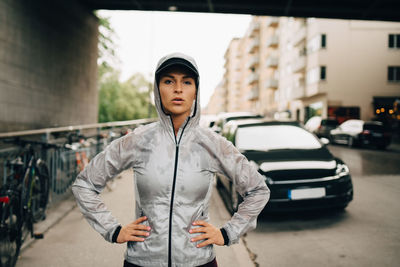 Portrait of confident female athlete in wet hooded jacket standing on sidewalk in city