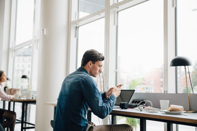 Young businessman using smart phone while sitting with laptop at desk in office